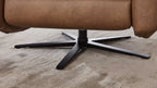 Eine braune Polster-Armlehne steht auf einem kunststoffbeschichteten, schwarzen Metallfuß mit einem fünfspitzigen Design auf einem hellen Teppich.