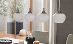 Vier hängende Lampen mit opalweißen Glaskörpern und metallenen Aufhängungen über einem Esstisch, im Hintergrund ein Pflanzenkübel.