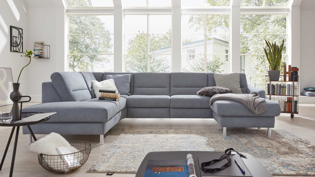 Graublaues Ecksofa mit Chaiselongue rechts, mehreren Kissen und Strickdecke vor großer Fensterfront, rechts Regal mit Pflanze und Büchern.