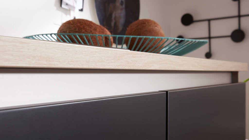Schrank mit dunkelgrauen Fronten und heller Holzplatte, darauf eine türkisfarbene Drahtschale mit zwei Kokosnüssen.