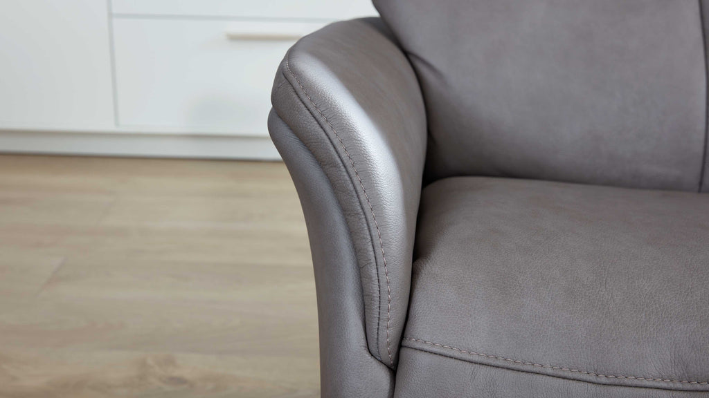 Graues Lederarmteil und Sitzfläche eines Sofas mit sichtbaren Nähten, im Hintergrund Holzboden und weiße Schrankfront.