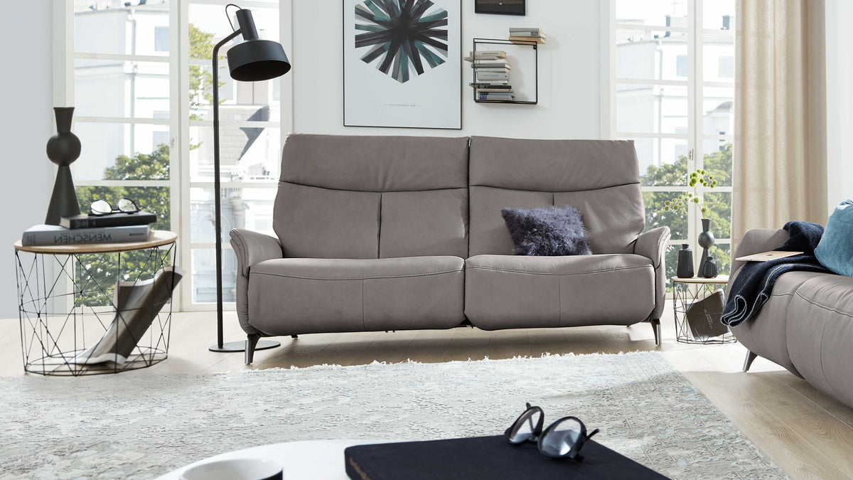 Grau bezogene Doppelsofa mit hohen Rückenlehnen und einem dunkelgrauen Fellkissen, flankiert von Stehlampe und Beistelltischen.