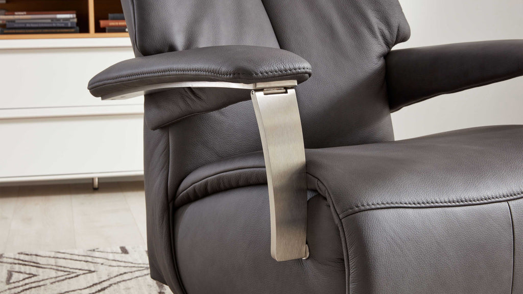 Dunkelgrauer Leder-Sesselsitz mit gepolstertem Armteil und gebürstetem Metallarmstütze, im Hintergrund weiße Kommode und Teppich.