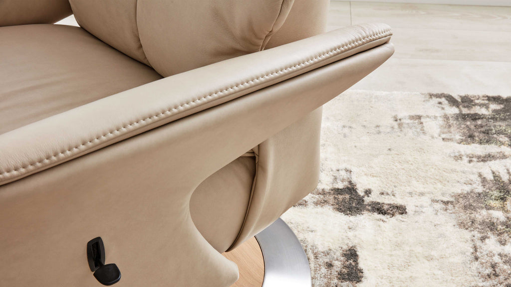 Nahaufnahme eines hellbeigen Leder-Sessels mit sichtbarer Ziernaht, schwarzem Hebel, rundem Metall-Holzfuß und Musterteppich.
