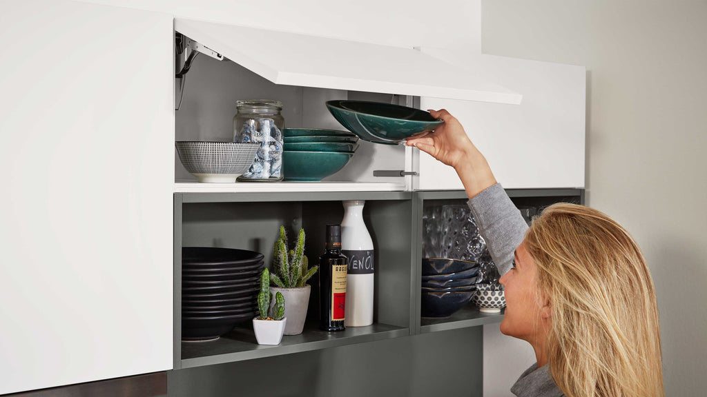 Hängeschrank mit aufgeklappter weißer Klappe, eine Frau entnimmt grüne Schalen; darunter Teller, Gläser, Flaschen und kleine Kakteen.
