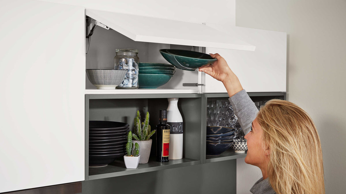 Hängeschrank mit aufgeklappter weißer Klappe, eine Frau entnimmt grüne Schalen; darunter Teller, Gläser, Flaschen und kleine Kakteen.