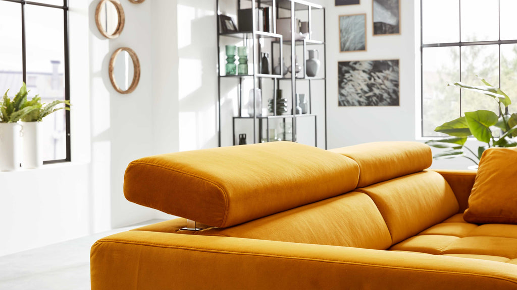 Senfgelbes Sofa mit verstellbarem Kopfteil und einzelnen Kissen, platziert in hellem Raum vor großen Fensterflächen.