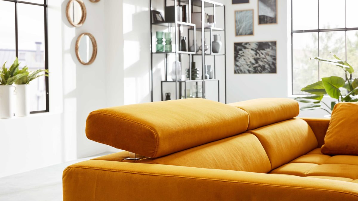 Senfgelbes Sofa mit verstellbarem Kopfteil und einzelnen Kissen, platziert in hellem Raum vor großen Fensterflächen.