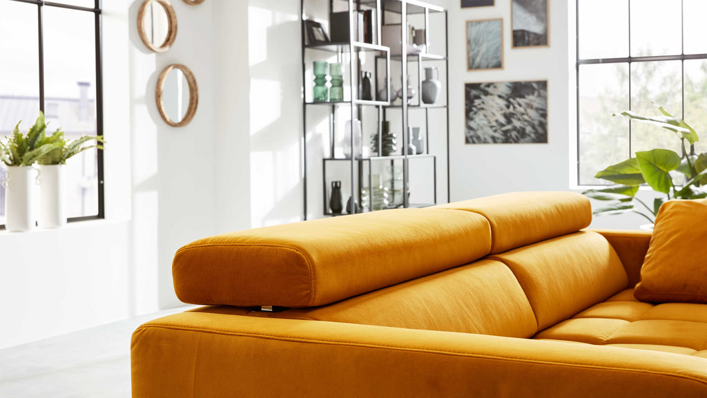 Ockerfarbenes Sofa mit verstellbarer Kopfstütze im Vordergrund, dahinter Regal, Fenster, Pflanzen und Wanddekorationen.