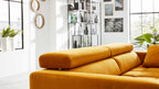 Ockerfarbenes Sofa mit verstellbarer Kopfstütze im Vordergrund, dahinter Regal, Fenster, Pflanzen und Wanddekorationen.