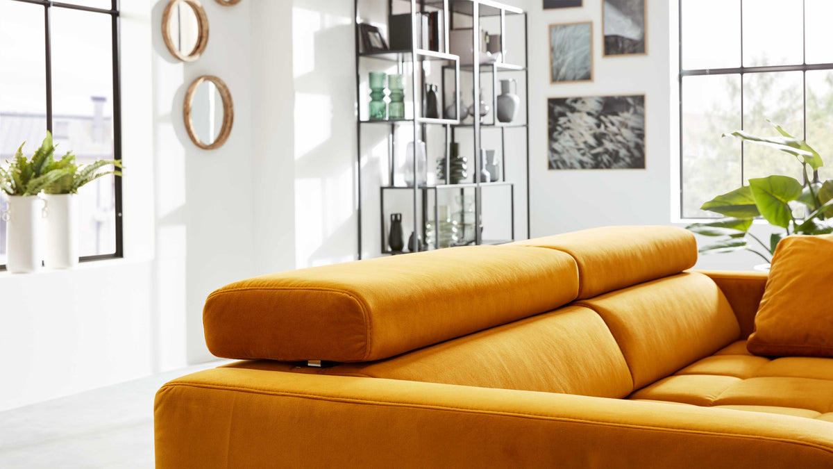Ockerfarbenes Sofa mit verstellbarer Kopfstütze im Vordergrund, dahinter Regal, Fenster, Pflanzen und Wanddekorationen.