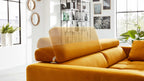 Oranger Ecksofa mit hochklappbarem Kopfteil, im hellen Wohnraum vor großen Gitterfenstern und Pflanzen.
