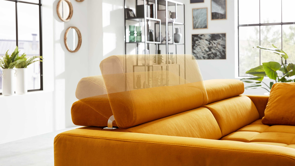 Oranger Ecksofa mit hochklappbarem Kopfteil, im hellen Wohnraum vor großen Gitterfenstern und Pflanzen.