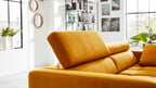Senfgelbes Sofa mit hochgestellter Kopfstütze in einem hellen Wohnzimmer mit Pflanzen, Regalen und mehreren Wandbildern.