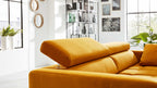 Senfgelbes Sofa mit hochgestellter Kopfstütze im Vordergrund, dahinter großes Fenster, Pflanzen und Regal.