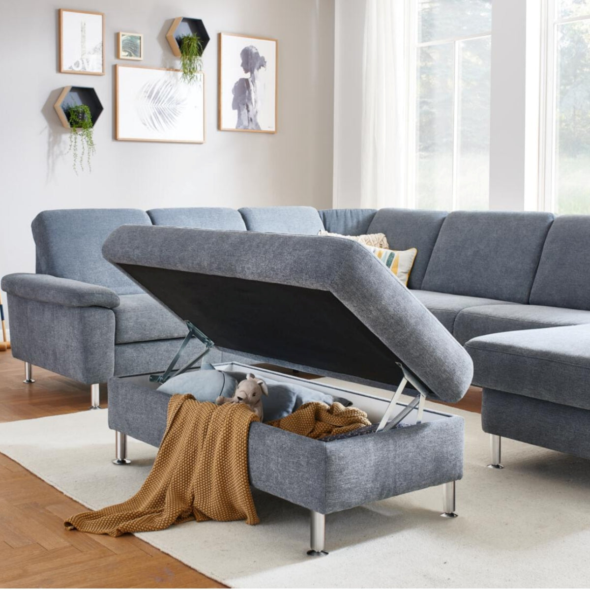 Ein grauer Modulmaster MM-ZE1129 XXL-Hocker von ZEHDENICK lässt sich aufklappen und gibt den Blick auf eine Decke und ein Spielzeug in einem modernen Wohnzimmer frei.