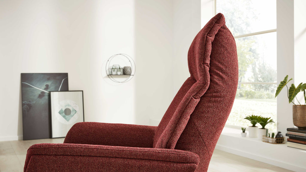 Roter Polstersessel mit hoher Lehne im Vordergrund, dahinter Fenster, Pflanzen und zwei gerahmte Bilder, an die Wand gelehnt.