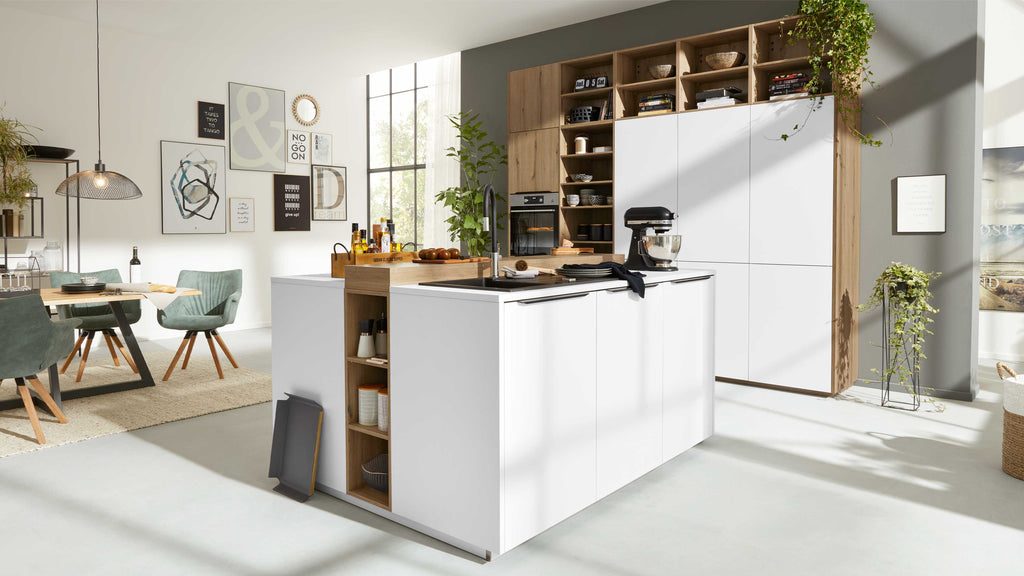 Moderne Küche mit einer großen Kochinsel in Weiß, einem Holzregal im Hintergrund, und einem Essbereich mit grünen Stühlen und einer Holztischplatte.