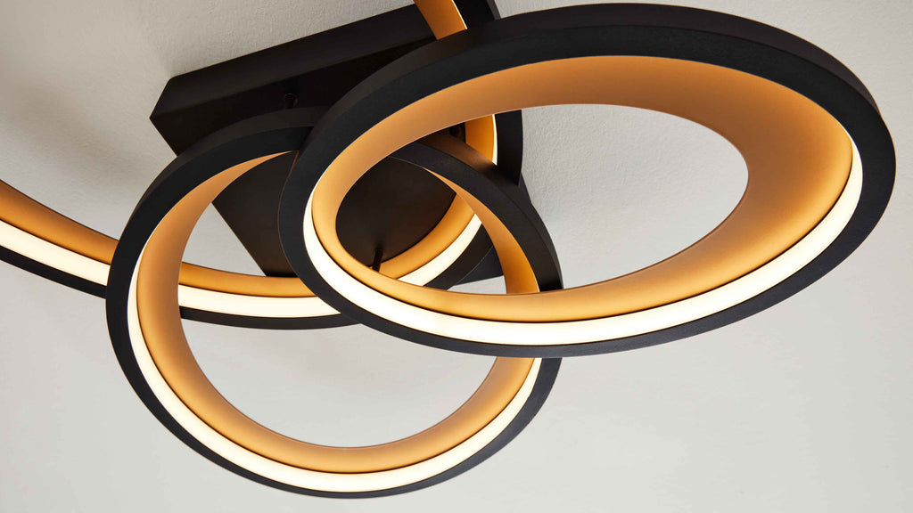 Deckenleuchte mit drei überlappenden schwarzen LED-Ringen, innen warm beleuchtet und an einer rechteckigen Deckenplatte montiert.