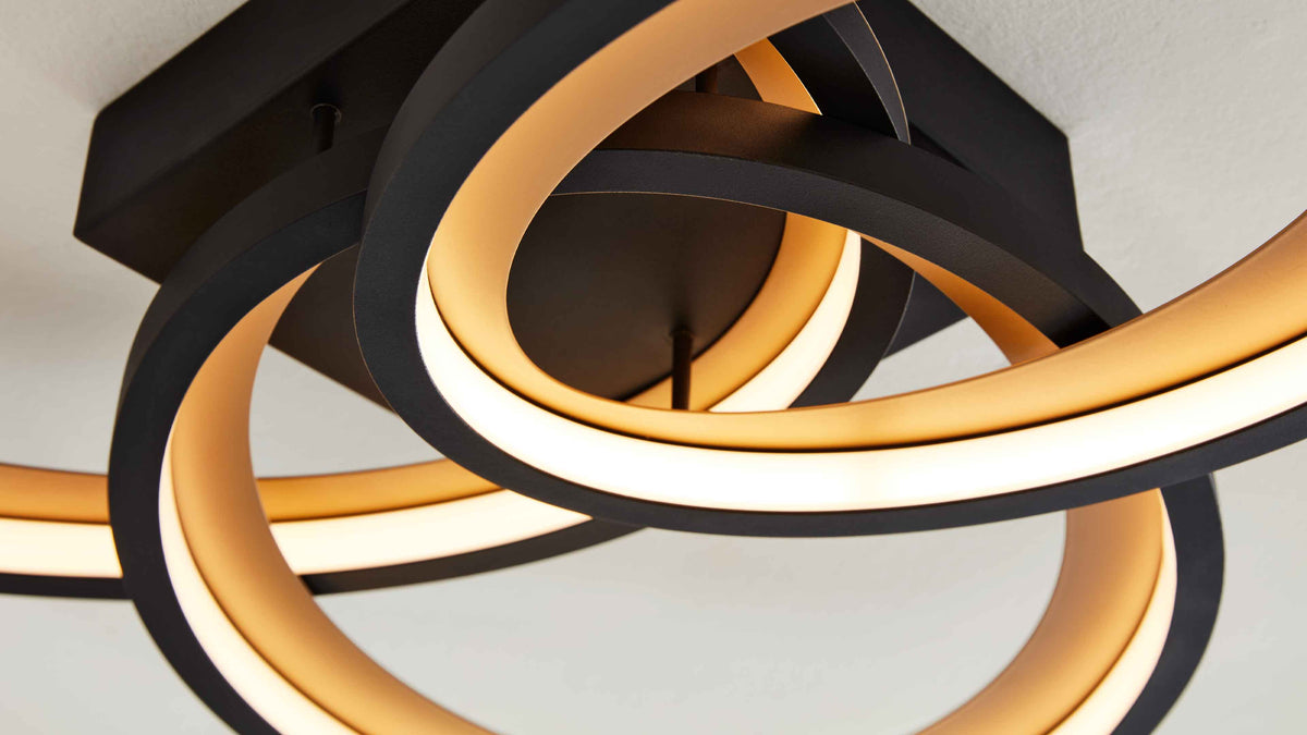 Schwarze Deckenleuchte mit ineinander verschlungenen Ringen und warmweiß leuchtenden LED-Streifen, befestigt an quadratischer Platte.