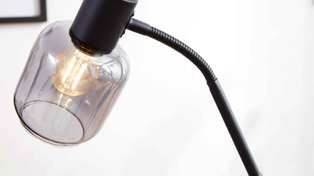 Schwarze Lampe mit flexiblem Hals, rauchfarbigem Glasschirm und sichtbarer Leuchtfaden-Glühbirne vor weißem Hintergrund.