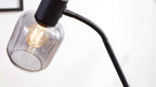Schwarze Lampe mit flexiblem Hals, rauchfarbigem Glasschirm und sichtbarer Leuchtfaden-Glühbirne vor weißem Hintergrund.