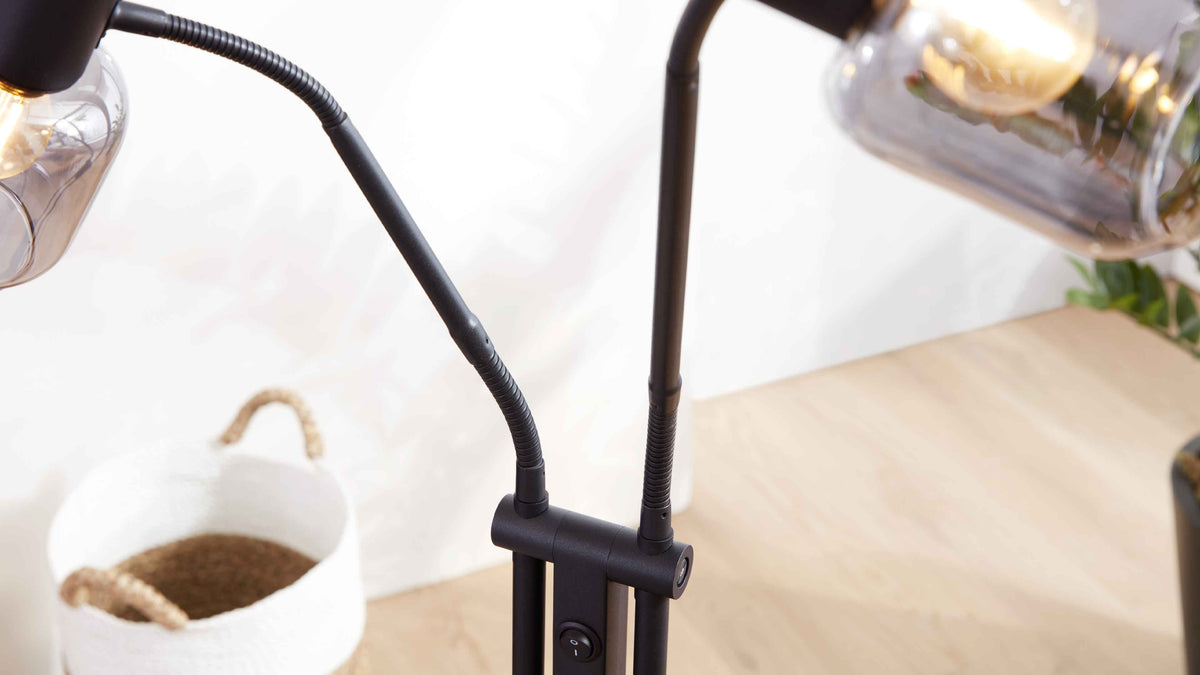 Schwarze Doppel-Leselampe mit zwei flexiblen Schwanenhälsen und rauchigen Glaskolben, auf hellem Holztisch neben weißem Korb.