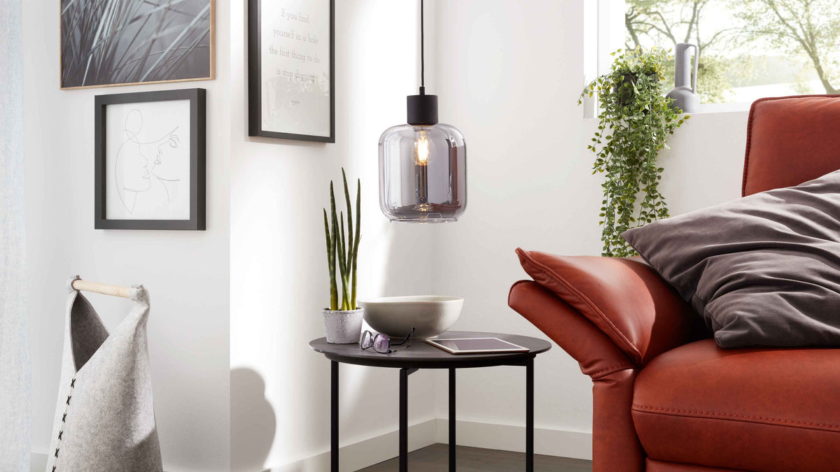 Rotes Sofa mit grauem Kissen, daneben runder schwarzer Beistelltisch mit Topfpflanze, Schale, Brille, Tablet und hängender Glaslampe.