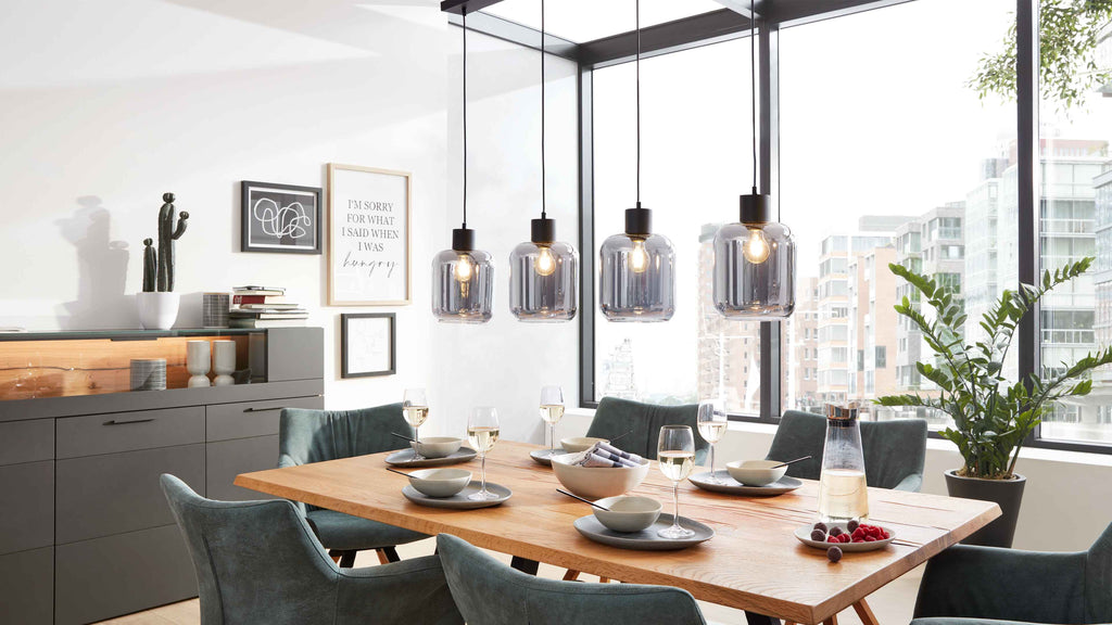 Massiver Holztisch mit Tellern, Gläsern und grünen Polsterstühlen, darüber vier transparente Hängelampen; Sideboard und Fenster.