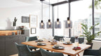 Massiver Holztisch mit Tellern, Gläsern und grünen Polsterstühlen, darüber vier transparente Hängelampen; Sideboard und Fenster.