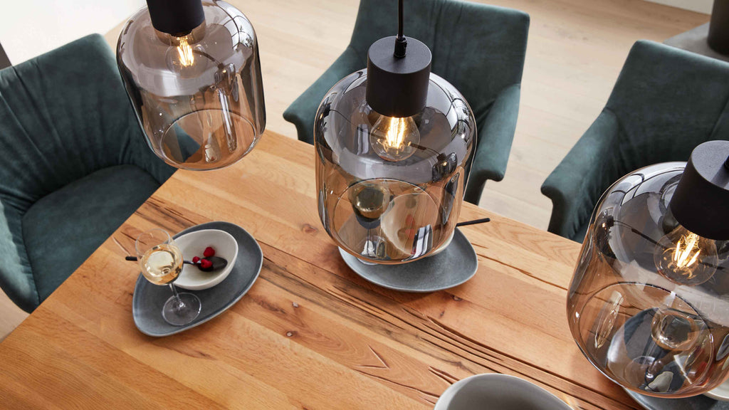 Große rauchglas-Pendelleuchte über Holztisch, dazu grüne Polsterstühle und Teller mit Weinglas und Schale.