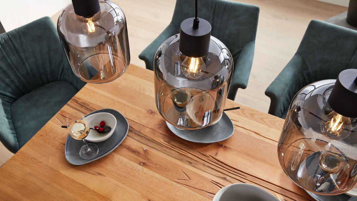 Große rauchglas-Pendelleuchte über Holztisch, dazu grüne Polsterstühle und Teller mit Weinglas und Schale.