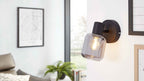 Wandlampe mit rundem dunklem Sockel und rauchfarbener Glasfassung mit leuchtender Birne, daneben Bilder, Fenster, Sofa und Pflanze.