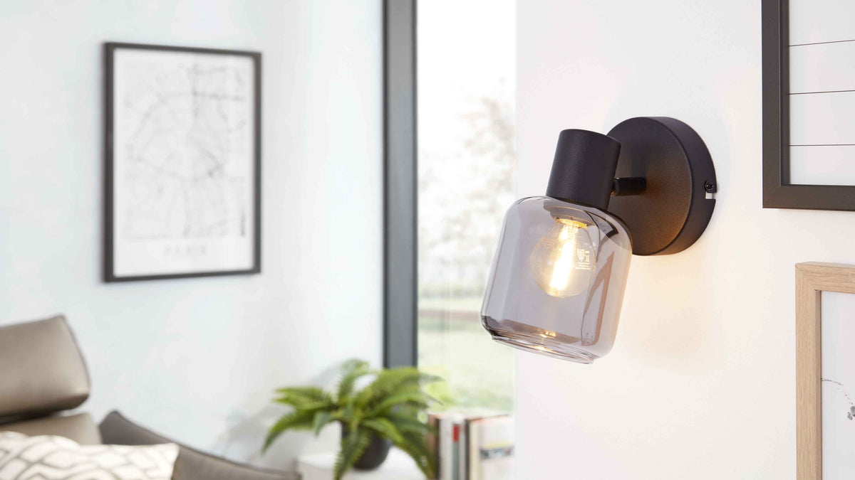 Wandlampe mit rundem dunklem Sockel und rauchfarbener Glasfassung mit leuchtender Birne, daneben Bilder, Fenster, Sofa und Pflanze.