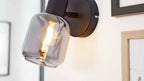 Wandlampe mit schwarzem Metallfuß und rauchfarbenem Glas-Schirm mit sichtbarer warm leuchtender Filamentbirne, daneben Holzbilderrahmen.