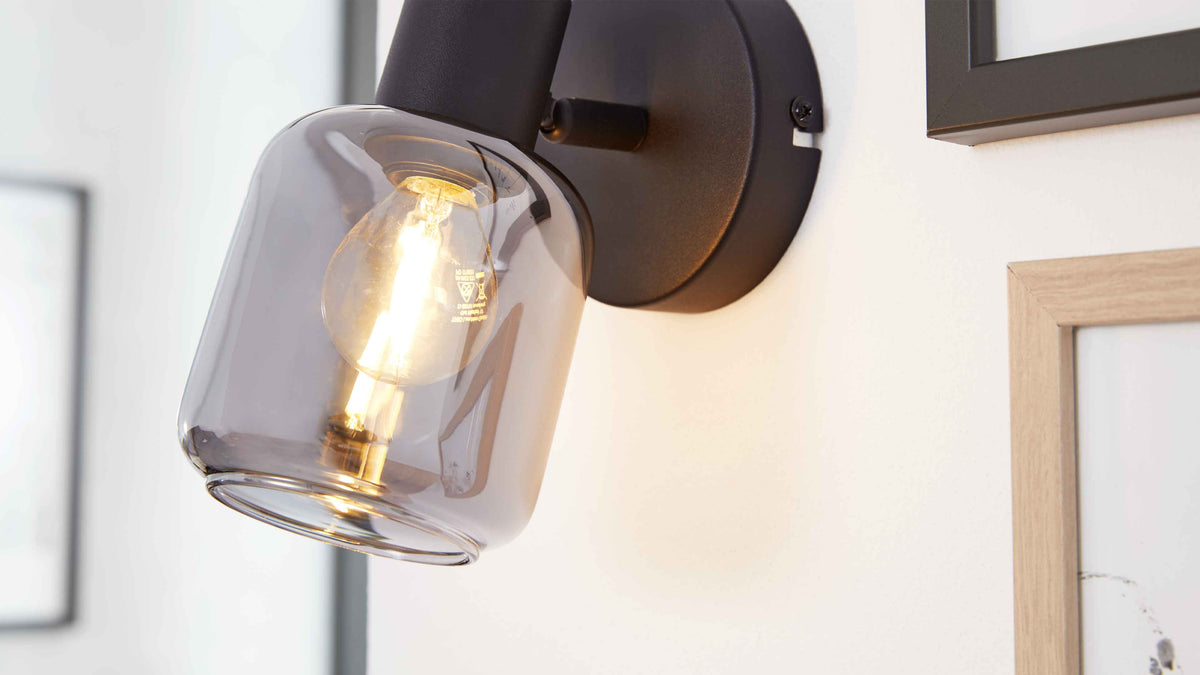 Wandlampe mit schwarzem Metallfuß und rauchfarbenem Glas-Schirm mit sichtbarer warm leuchtender Filamentbirne, daneben Holzbilderrahmen.