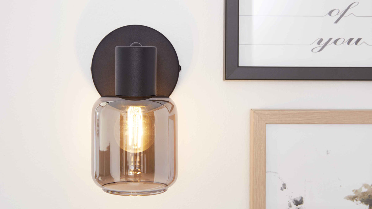 Wandleuchte mit rauchfarbenem Glas und sichtbarer Leuchtfadenlampe an schwarzer Rundhalterung an weißer Wand, rechts zwei Bilderrahmen.