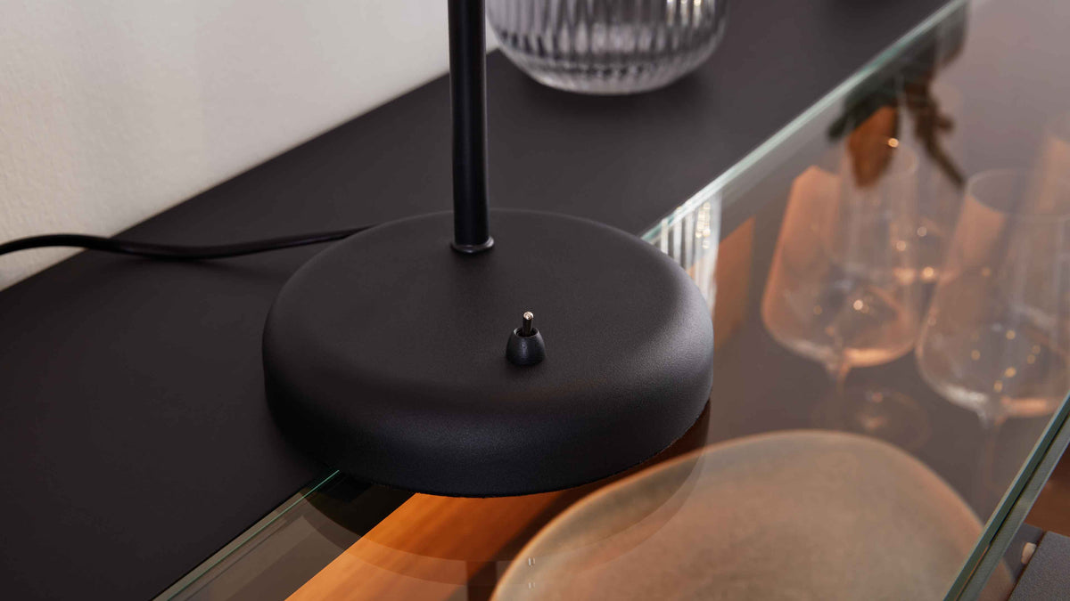 Schwarze runde Lampenfuß mit kleinem Kippschalter auf dunkler Tischplatte, Kabel sichtbar, Glasspiegelung und Weingläser im Hintergrund.