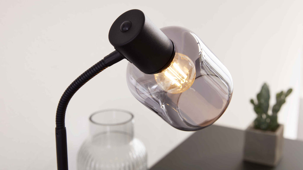 Schwarze Schreibtischlampe mit flexiblem Hals, Glasfassung und leuchtender Filamentbirne auf einem Tisch neben Glasvase und Kaktus.