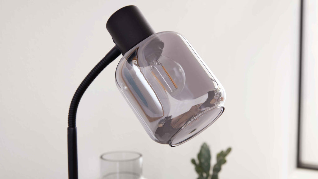 Schwarze Lampe mit flexiblem Hals und rauchigem Glasschirm, sichtbare Filament-Glühbirne; im Hintergrund Glasvase und grüne Zimmerpflanze.