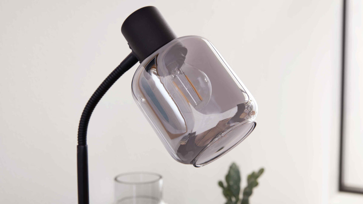 Schwarze Lampe mit flexiblem Hals und rauchigem Glasschirm, sichtbare Filament-Glühbirne; im Hintergrund Glasvase und grüne Zimmerpflanze.