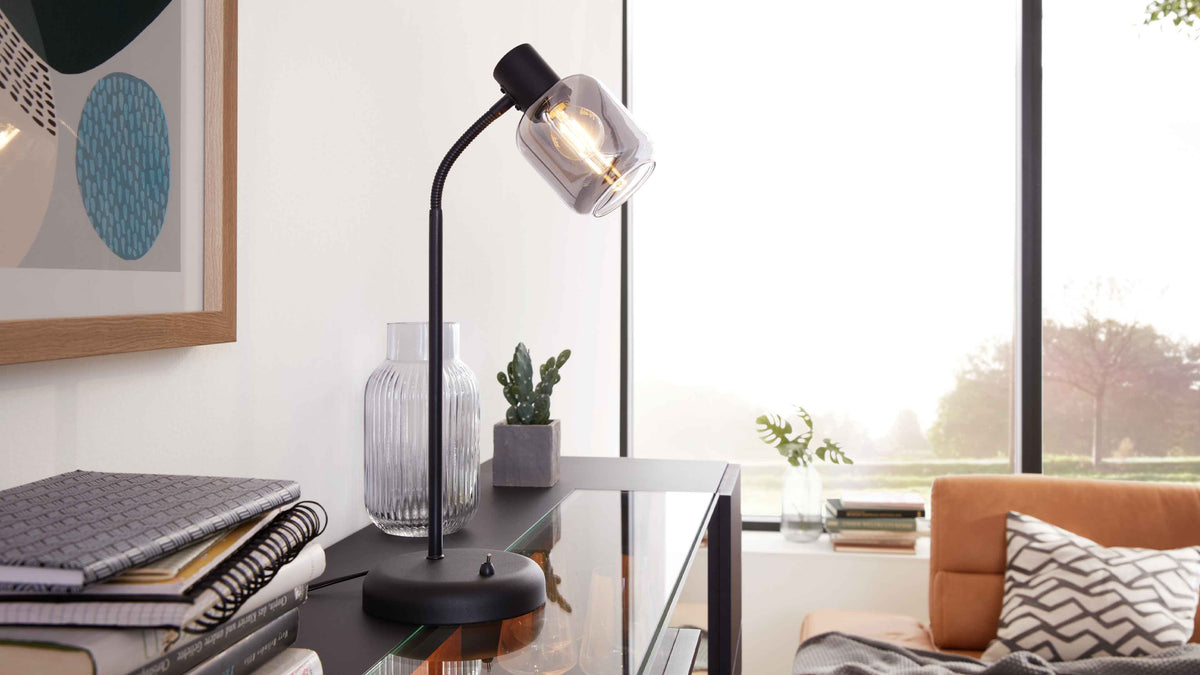 Schwarze Tischlampe mit rauchglaslampenschirm auf schwarzem Glas-Tisch, daneben Vasen, Kaktus und orange Couch mit Kissen.