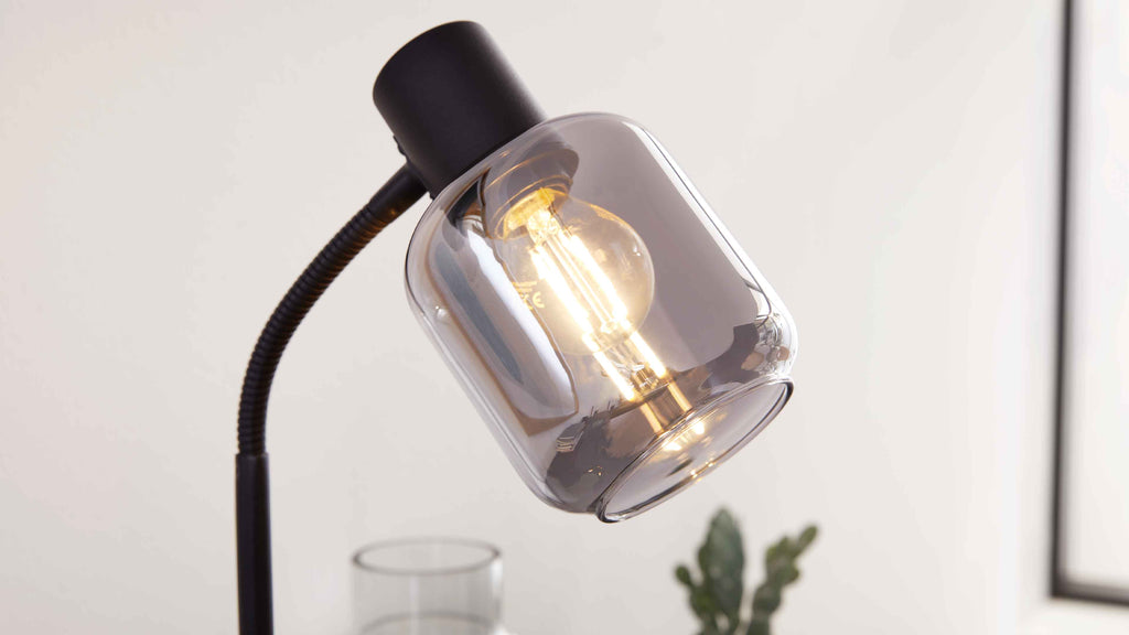 Schwarze Lese- oder Tischlampe mit flexiblem, gewölbtem Arm und rauchfarbenem Glasschirm, darin leuchtende Birne sichtbar.