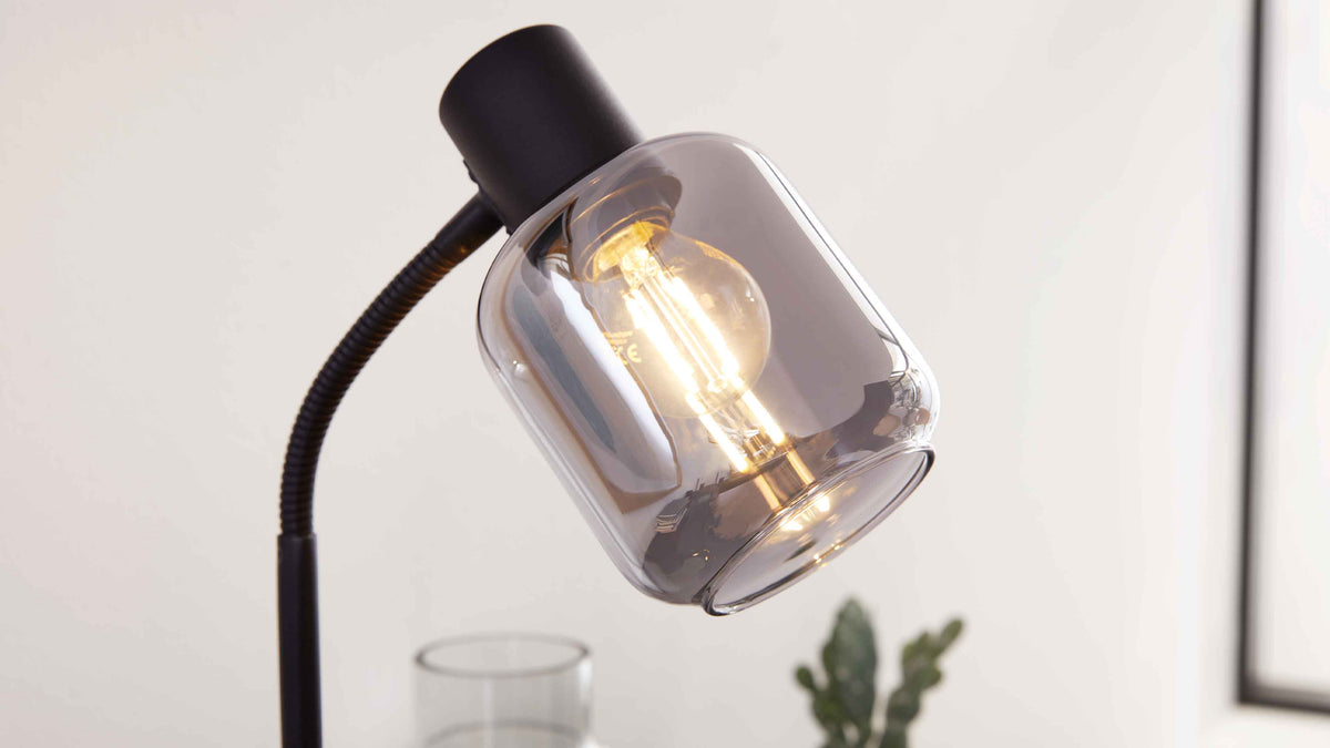 Schwarze Lese- oder Tischlampe mit flexiblem, gewölbtem Arm und rauchfarbenem Glasschirm, darin leuchtende Birne sichtbar.
