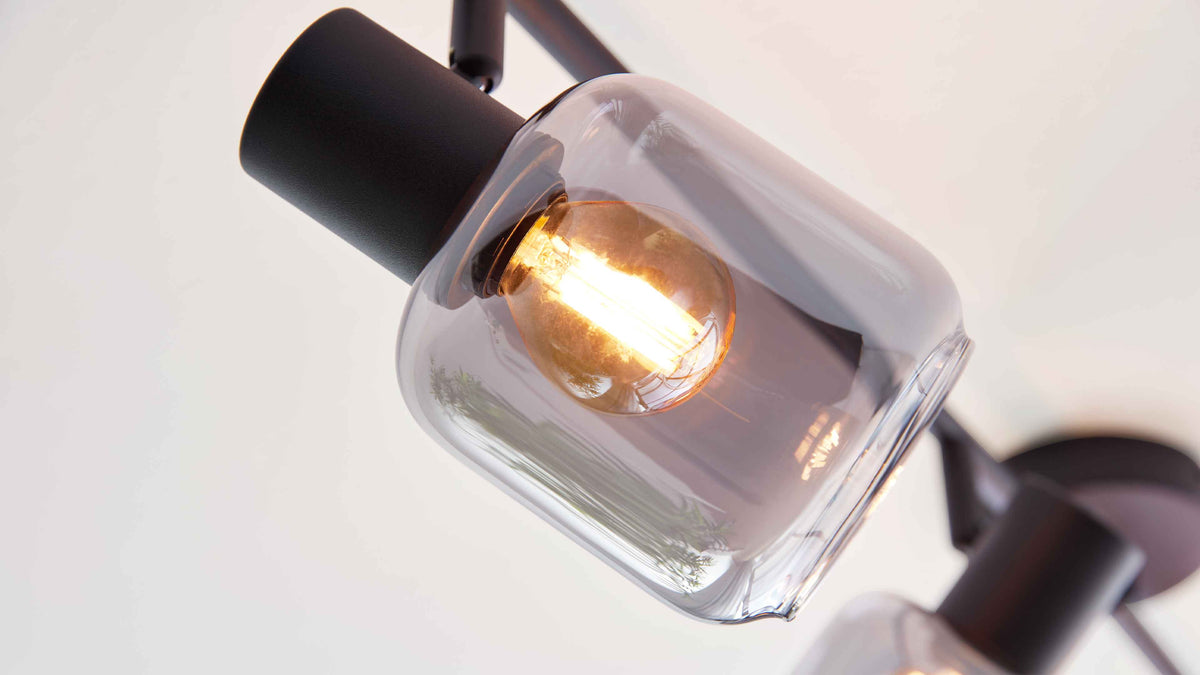 Nahaufnahme einer Deckenleuchte mit schwarzem Metallhalter und klarem, abgerundetem Glasschirm, darin leuchtende Filamentbirne.