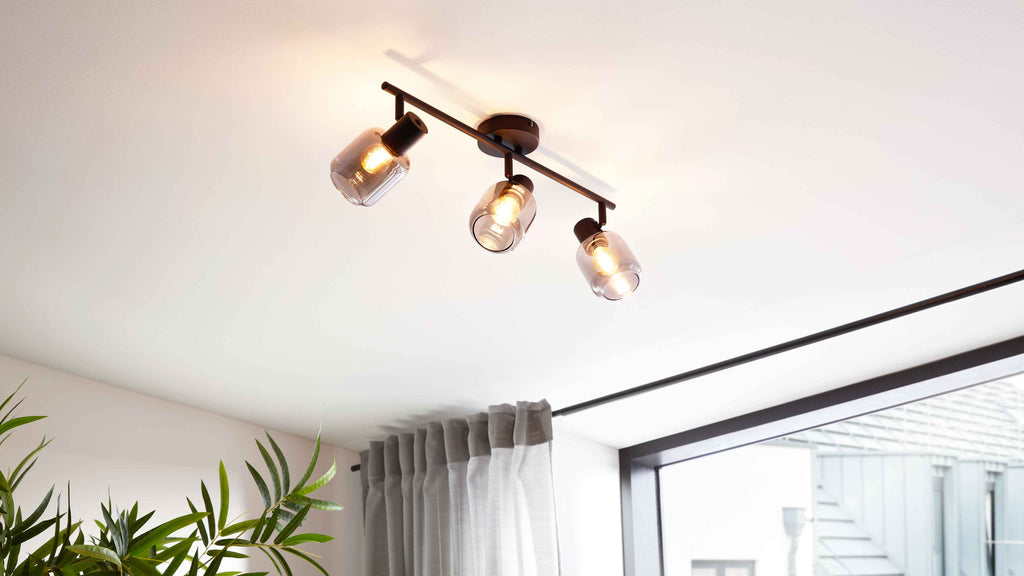 Dreiflammige Deckenleuchte mit dunklem Metallstab und rauchigen Glasglocken, daneben Gardine an Vorhangstange und Fenster.