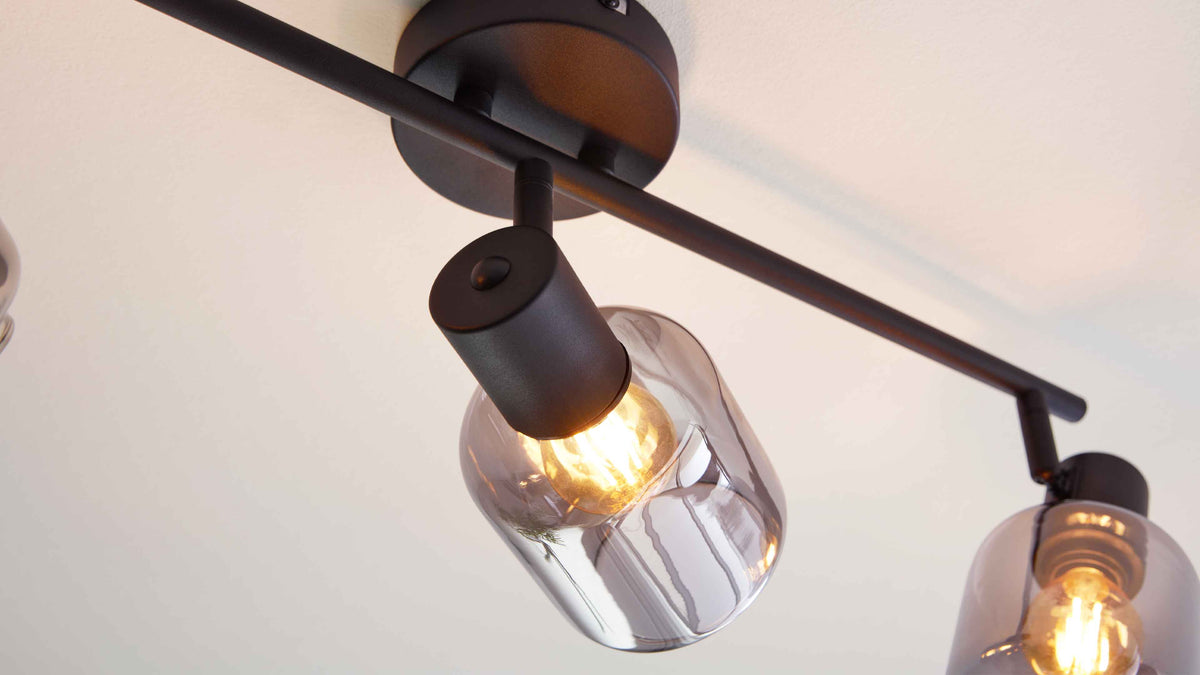 Deckeleuchte mit schwarzem Metallsteg und zwei schwenkbaren Fassungen, eine mit klarem Glaszylinder und leuchtender Filamentlampe.