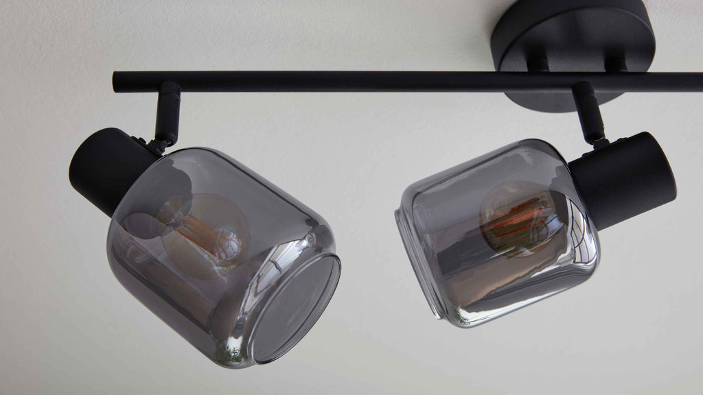 Deckenleuchte mit schwarzem Metallträger und zwei drehbaren Fassungen mit rauchgrauen Glaszylindern und sichtbaren Filamentlampen.