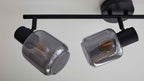 Deckenleuchte mit schwarzem Metallträger und zwei drehbaren Fassungen mit rauchgrauen Glaszylindern und sichtbaren Filamentlampen.