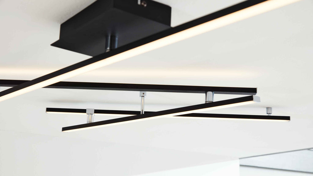 Deckenleuchte mit mehreren sich kreuzenden schwarzen LED-Linienstäben, an silbernen Halterungen an weißer Decke montiert.